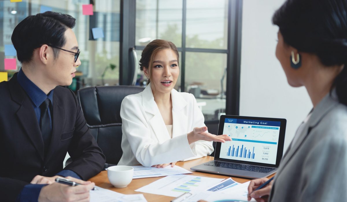 Business Meeting Discussing Marketing Goals and Financial Plans Group of Asian business professionals in a modern office setting discussing marketing goals and financial plans, with charts and graphs displayed on a laptop.